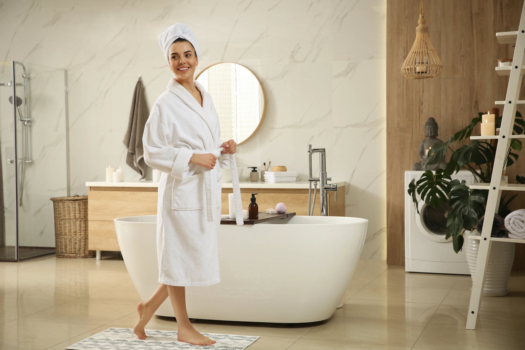 Eine Frau in einem weißen Bademantel steht lächelnd in einem modernen Badezimmer. Im Hintergrund ist ein runder Spiegel über einem Waschbecken zu sehen, daneben hängt ein graues Handtuch. Im Vordergrund befindet sich eine freistehende Badewanne mit einem Holzbrett, auf dem Badeutensilien liegen. Die Umgebung ist hell und stilvoll eingerichtet, mit Pflanzen, Kerzen und dekorativen Elementen.