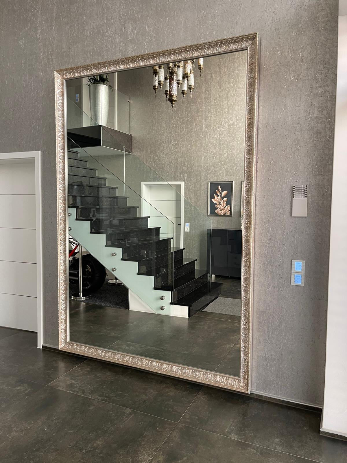 Großer Wandspiegel mit dekorativem Rahmen im Flur, spiegelt eine Treppe mit Glasgeländer und Kronleuchter.