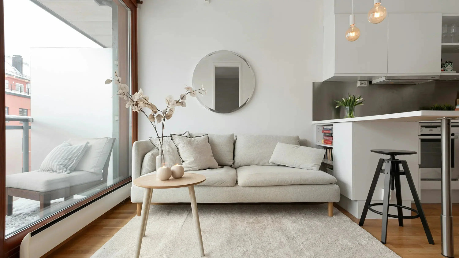 Helles, modern eingerichtetes Wohnzimmer mit einem großen runden Wandspiegel über einem hellen Sofa, stilvollem Coffee-Table und angrenzender offener Küche – ideal zur Präsentation hochwertiger runder Spiegel für Wohnräume.