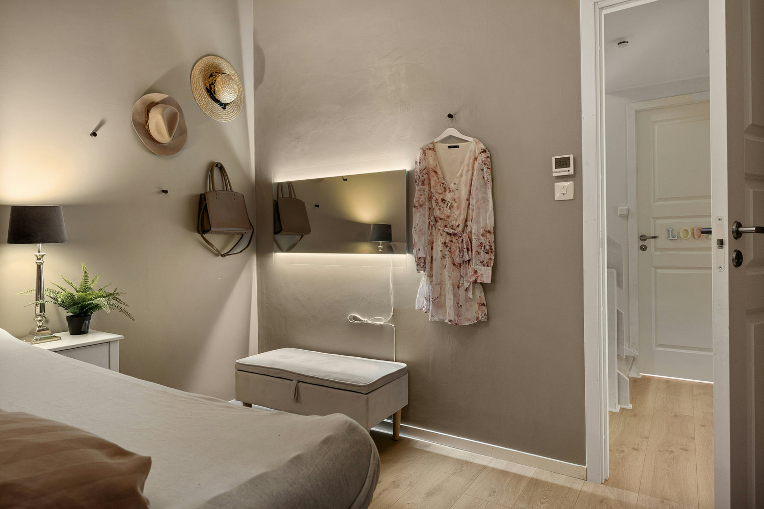 Kleiner, gemütlicher Ankleidebereich mit beleuchtetem Wandspiegel, einer gepolsterten Sitzbank, aufgehängtem Kleid, Hüten und Tasche an der Wand sowie Blick in einen angrenzenden Flur.