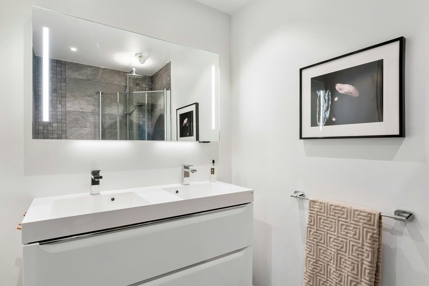 Modernes Badezimmer mit weißem Doppelwaschbecken, großem LED-beleuchtetem Spiegel, grauer gefliester Duschkabine und wandhängem Handtuchhalter mit gemustertem Handtuch.