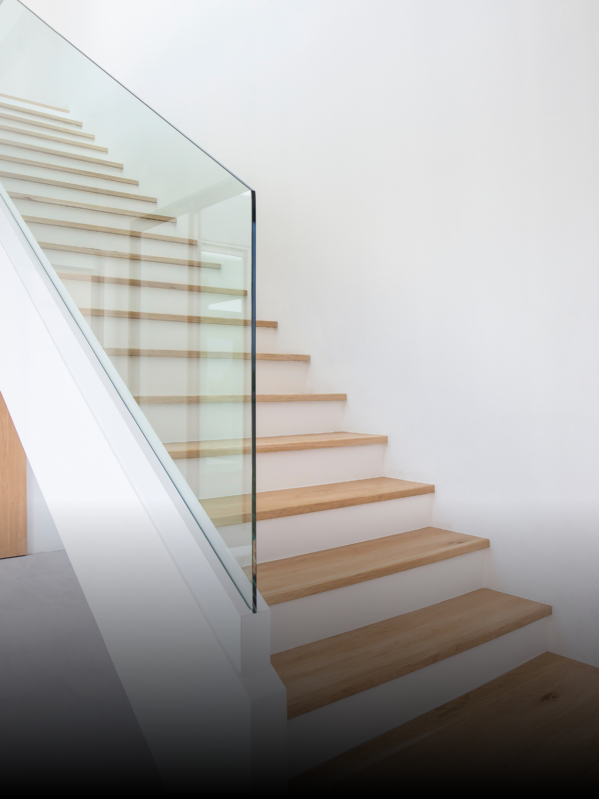 Eine moderne Innentreppe aus hellem Holz mit einem seitlichen Geländer aus VSG-Glas. Die klare Glasfläche verläuft entlang der Treppe und bildet einen eleganten Kontrast zu den weißen Wänden und den natürlichen Holzstufen.