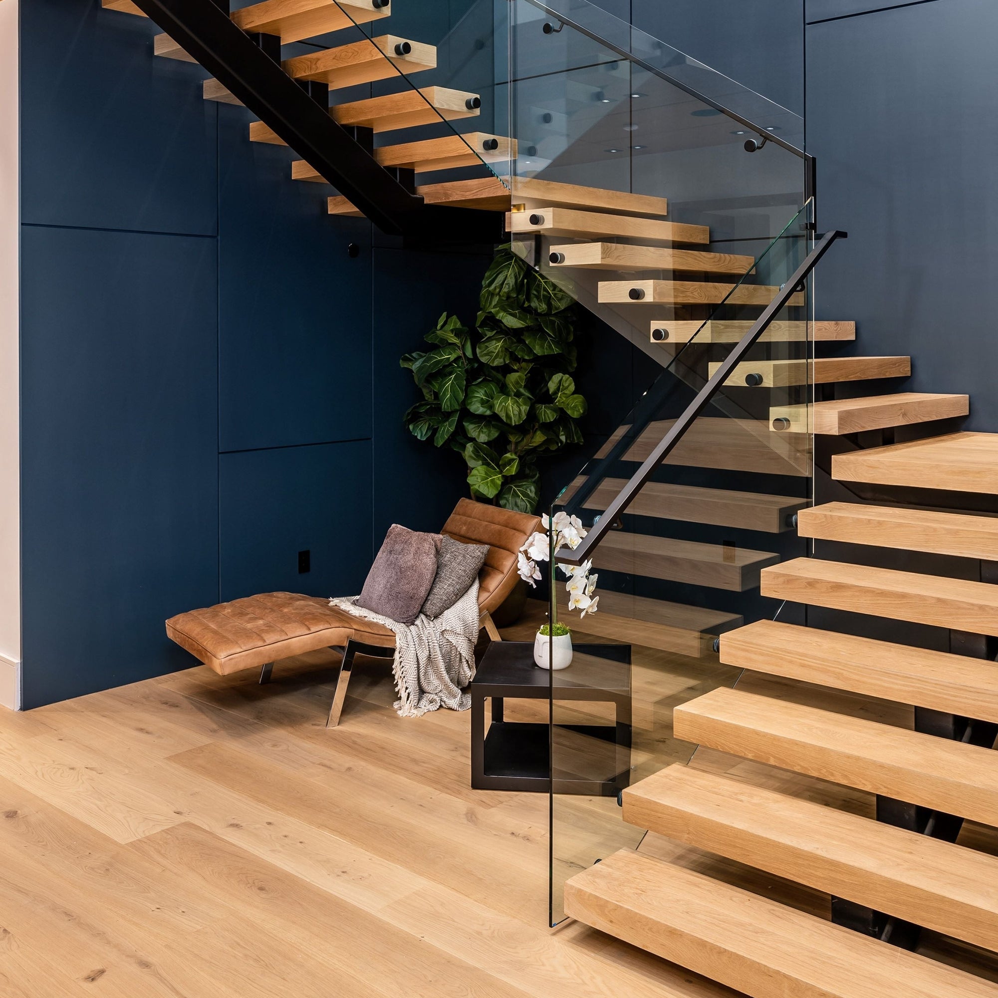Moderne Treppe aus hellem Holz mit Glasgeländer, darunter ein kleiner Sitzbereich mit Lederstuhl, Dekokissen und einer grünen Pflanze.