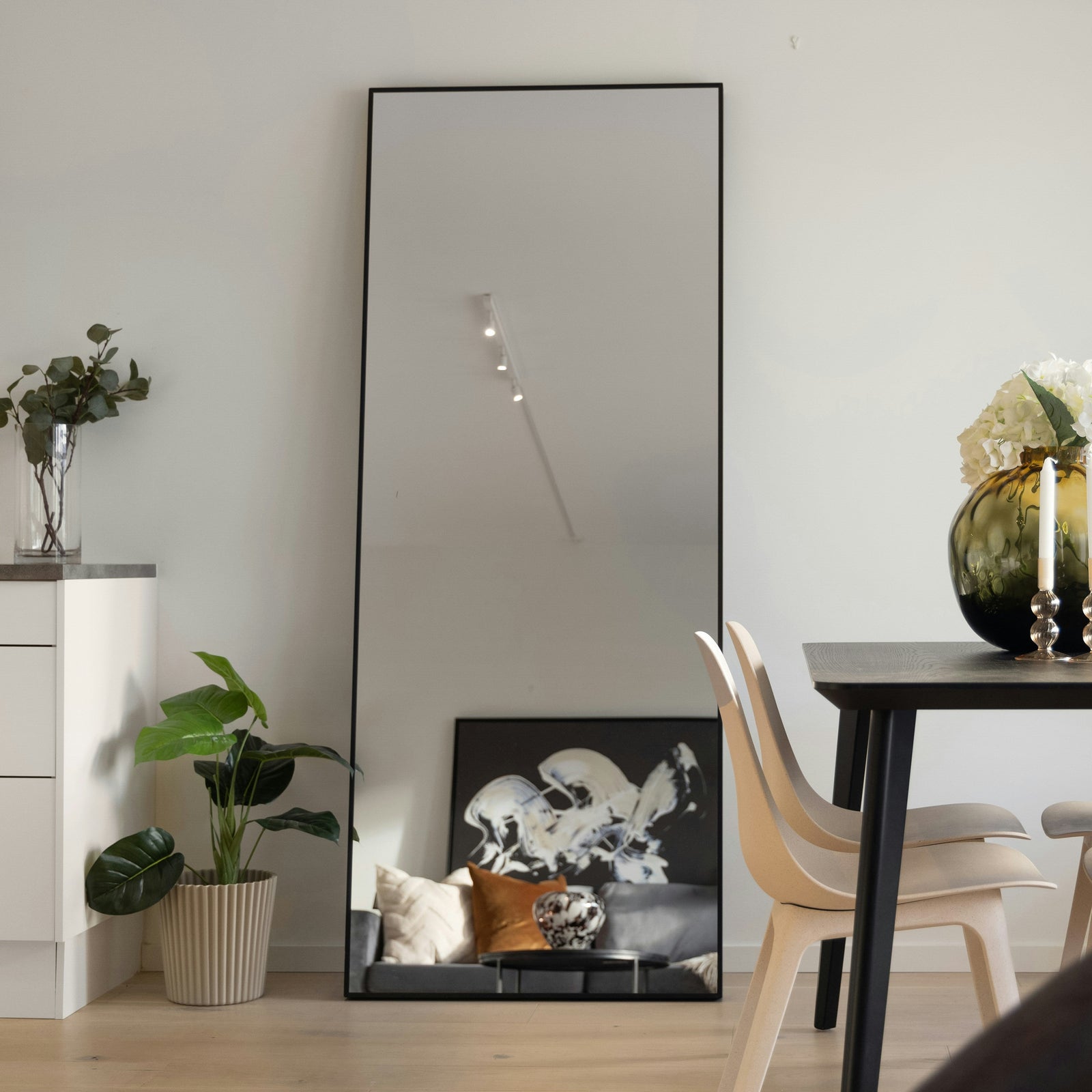Großer rechteckiger Standspiegel von Spiegelmax mit schwarzem Rahmen, der ein stilvoll eingerichtetes Wohnzimmer mit Kunstbild, Sofa und Pflanzen reflektiert. Der Spiegel steht an einer weißen Wand neben Esstisch und Dekoration.
