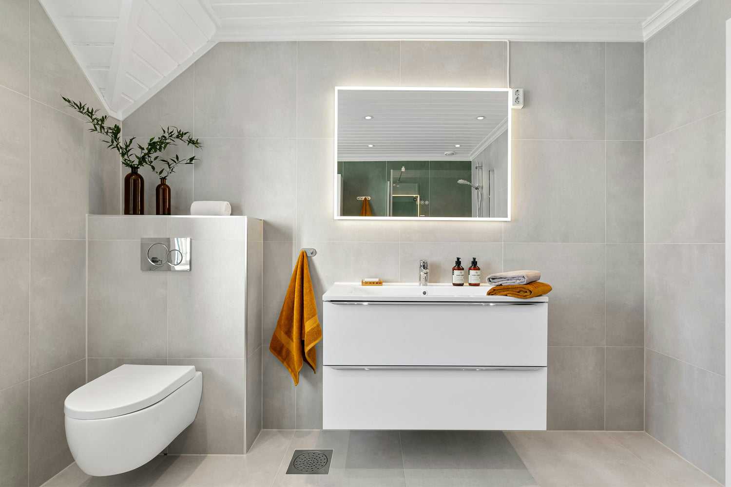 Ein helles, modern eingerichtetes Badezimmer mit großen grauen Wandfliesen, einem rechteckigen LED-Spiegel über einem weißen Waschtischunterschrank und einem wandhängenden WC. Auf dem Waschtisch stehen Pflegeprodukte und gefaltete Handtücher, daneben hängt ein senfgelbes Handtuch. Die Dusche im Hintergrund ist durch die Spiegelung sichtbar. Zwei dekorative braune Vasen mit Zweigen stehen auf einer kleinen Ablage neben dem WC.