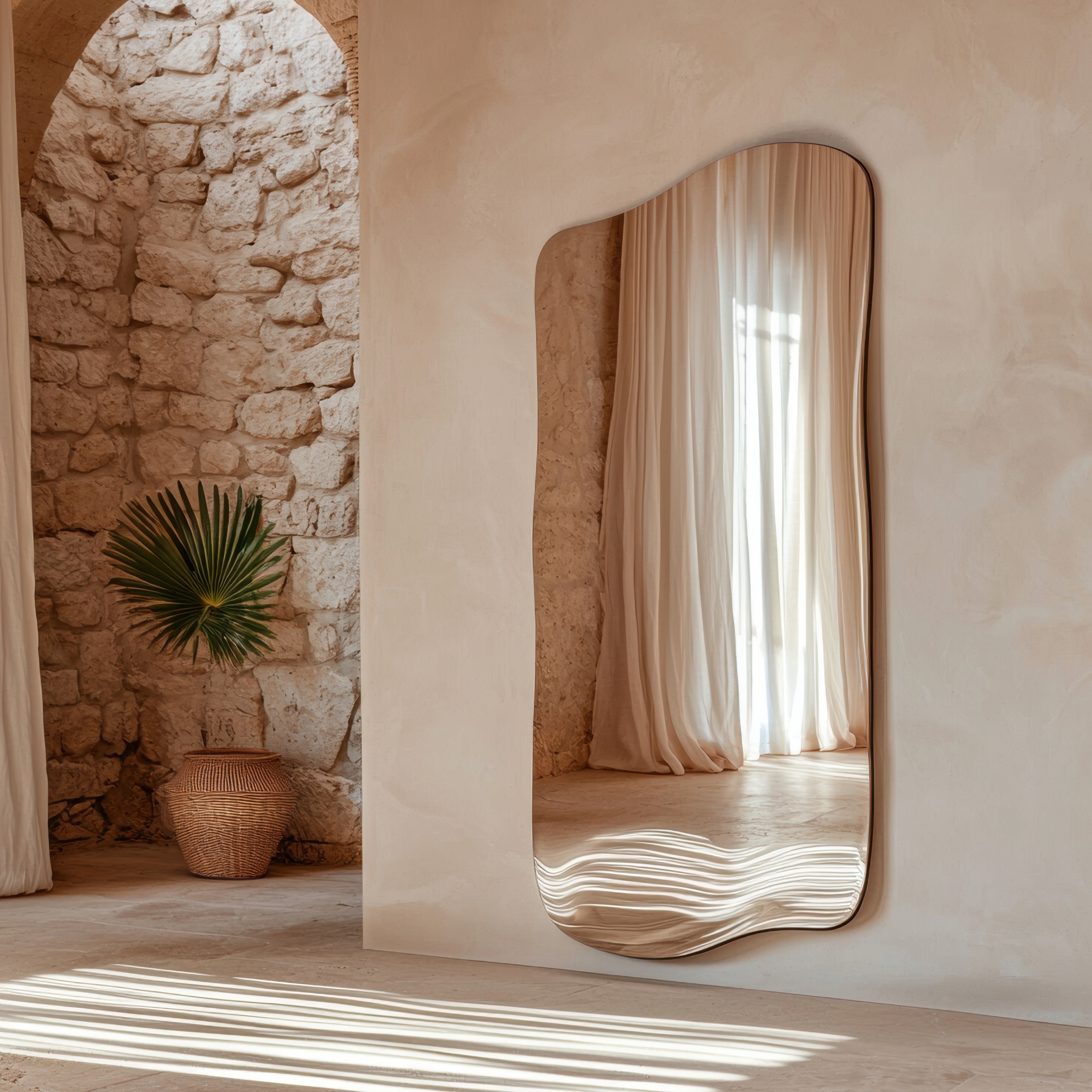 Ein organisch geformter Designspiegel von Spiegelmax hängt an einer cremefarbenen Wand in einem mediterranen Raum mit Natursteinmauer. Der Spiegel reflektiert sanft fallende Vorhänge und weiches Licht, während ein geflochtener Korb und ein grünes Palmenblatt eine natürliche, warme Atmosphäre schaffen.