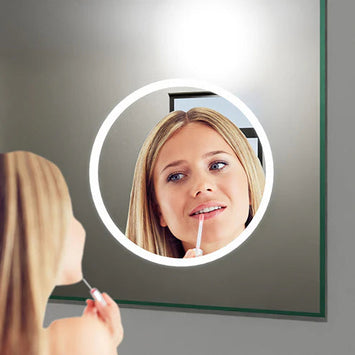 Frau trägt Lippenpflege auf und betrachtet sich in einem runden, beleuchteten Kosmetikspiegel, der auf einem großen Badezimmerspiegel montiert ist.