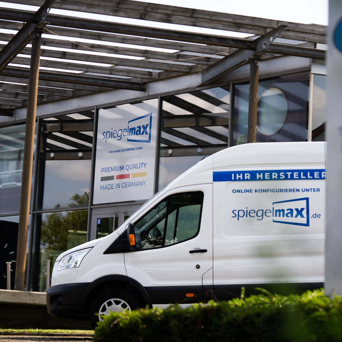 Das Firmengebäude von Spiegelmax mit großem Markenlogo und dem Hinweis „Premium Quality – Made in Germany“. Davor steht ein weißer Spiegelmax-Lieferwagen mit blauem Branding und dem Hinweis „Ihr Hersteller – Online konfigurieren unter spiegelmax.de“. Die Szene zeigt den Eingangsbereich des Unternehmens unter einer modernen Glas- und Holzüberdachung.