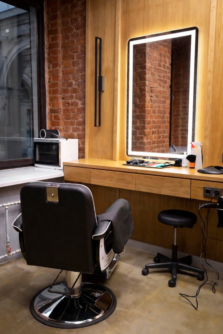 Moderner Barbershop Stylingplatz mit beleuchtetem Friseurspiegel, Holzablage, Barberstuhl und industrieller Backsteinwand