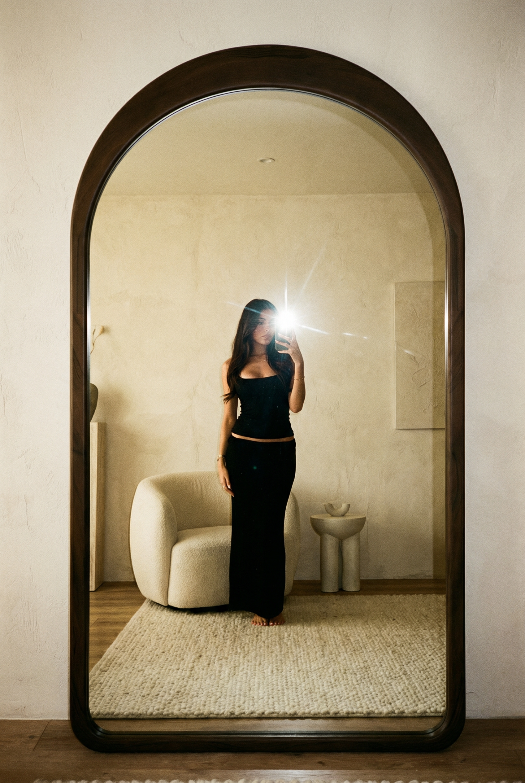 Bogenförmiger Standspiegel mit dunklem Rahmen in einem minimalistischen Raum, Person im Spiegel fotografiert sich.