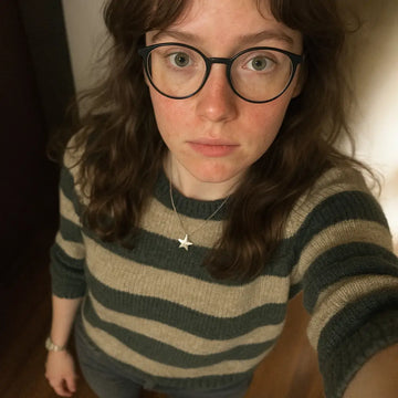 Junge Frau mit langen, braunen Haaren, runder Brille und ernstem Blick macht ein Selfie von oben. Sie trägt einen gestreiften Pullover in Beige und Dunkelgrün, eine Stern-Halskette und eine silberne Uhr. Die Aufnahme wirkt spontan und natürlich, gemacht in einem Innenraum mit warmem Licht