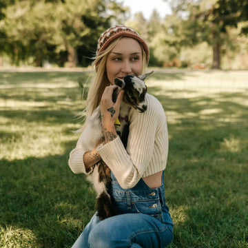 Junge Frau mit blonden Haaren und Mütze sitzt im Garten und hält liebevoll ein kleines Zicklein im Arm. Sie trägt einen cremefarbenen Pullover und eine Latzhose, die Szene wirkt natürlich und entspannt