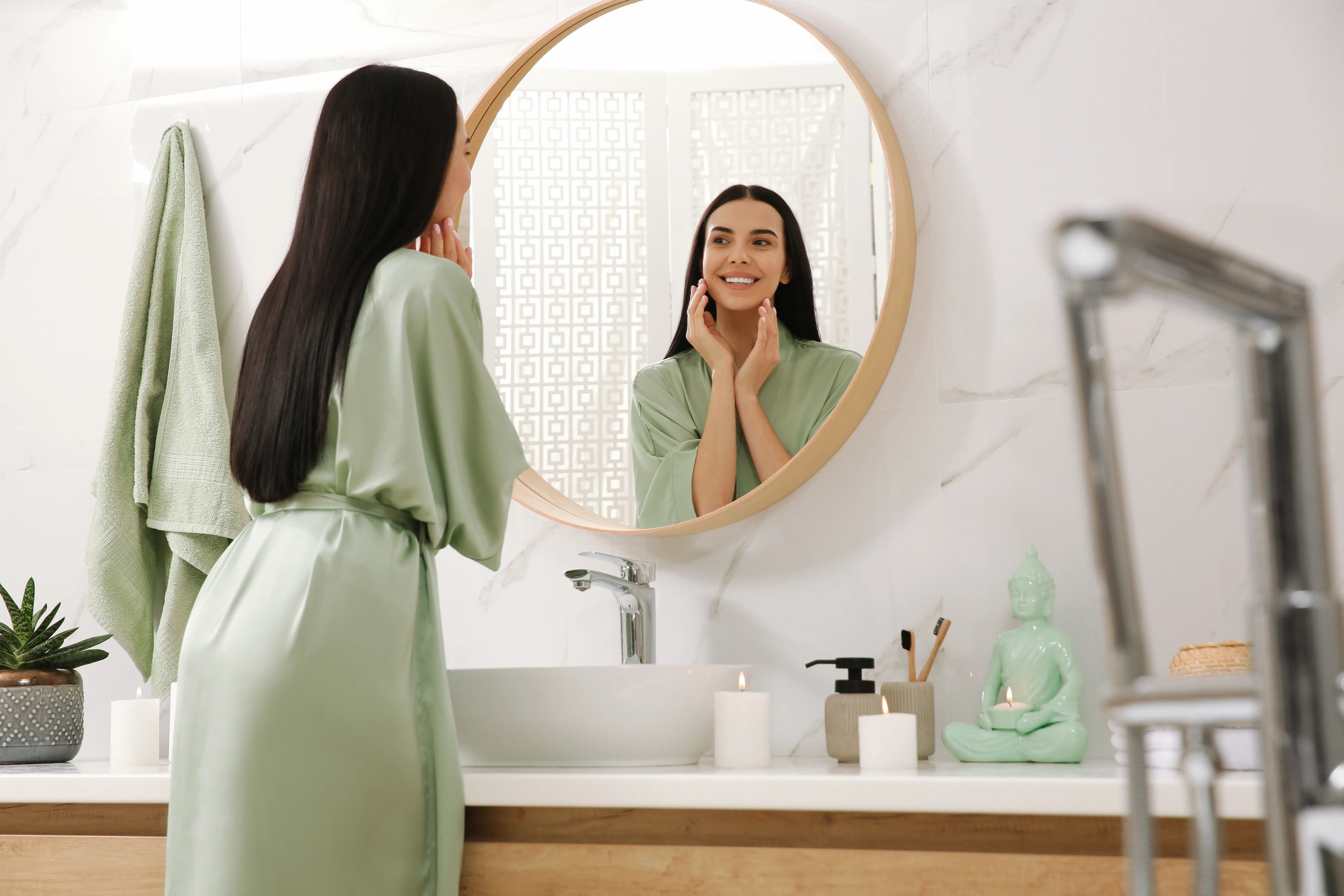 Eine Frau in einem mintgrünen Seidenbademantel steht vor einem runden Spiegel und berührt ihr Gesicht, während sie sich im Spiegelbild betrachtet. Das Badezimmer wirkt hell, ruhig und ist mit dezenten Accessoires und einem Buddha-Dekor gestaltet.