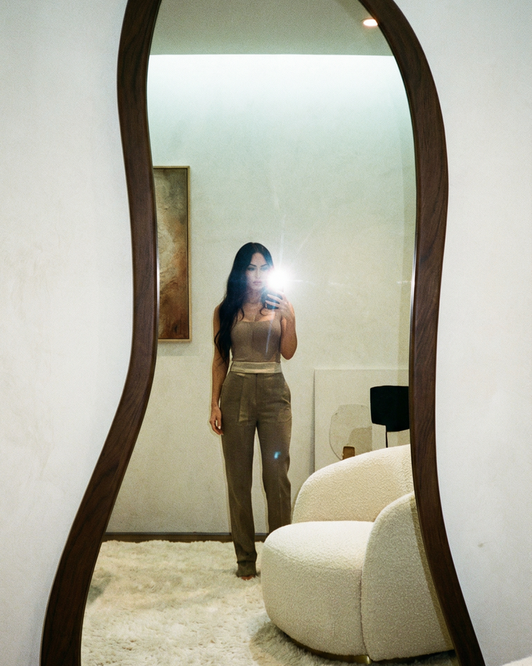 Ganzkörper-Spiegelselfie einer Frau in einem stilvollen, hellen Raum mit neutralem Interieur und cremefarbenem Sessel.